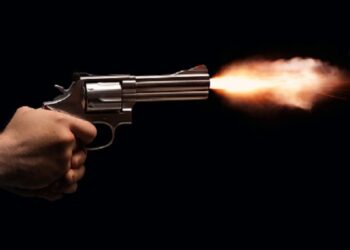 A revolver held by two anonymous hands is fired on a black background.
